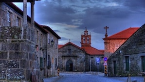 A cobblestone street with historic stone buildings, a church with a bell tower, and a parking sign. - Casa Aldeia da Lapa (Sernancelhe)