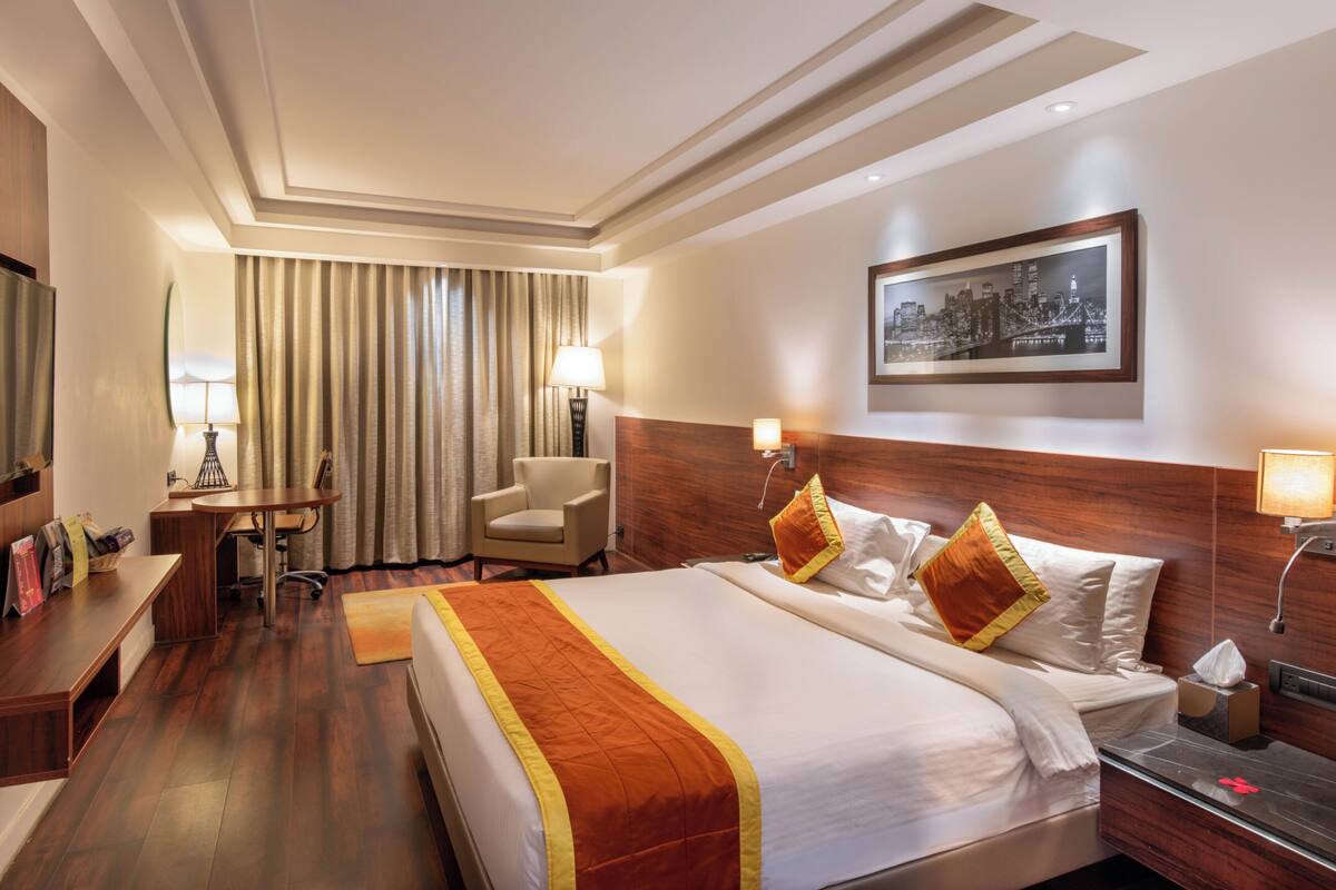 A hotel room with a large bed, a desk, a chair, a desk lamp, a bedside table, a nightstand, and a framed cityscape picture on the wall.