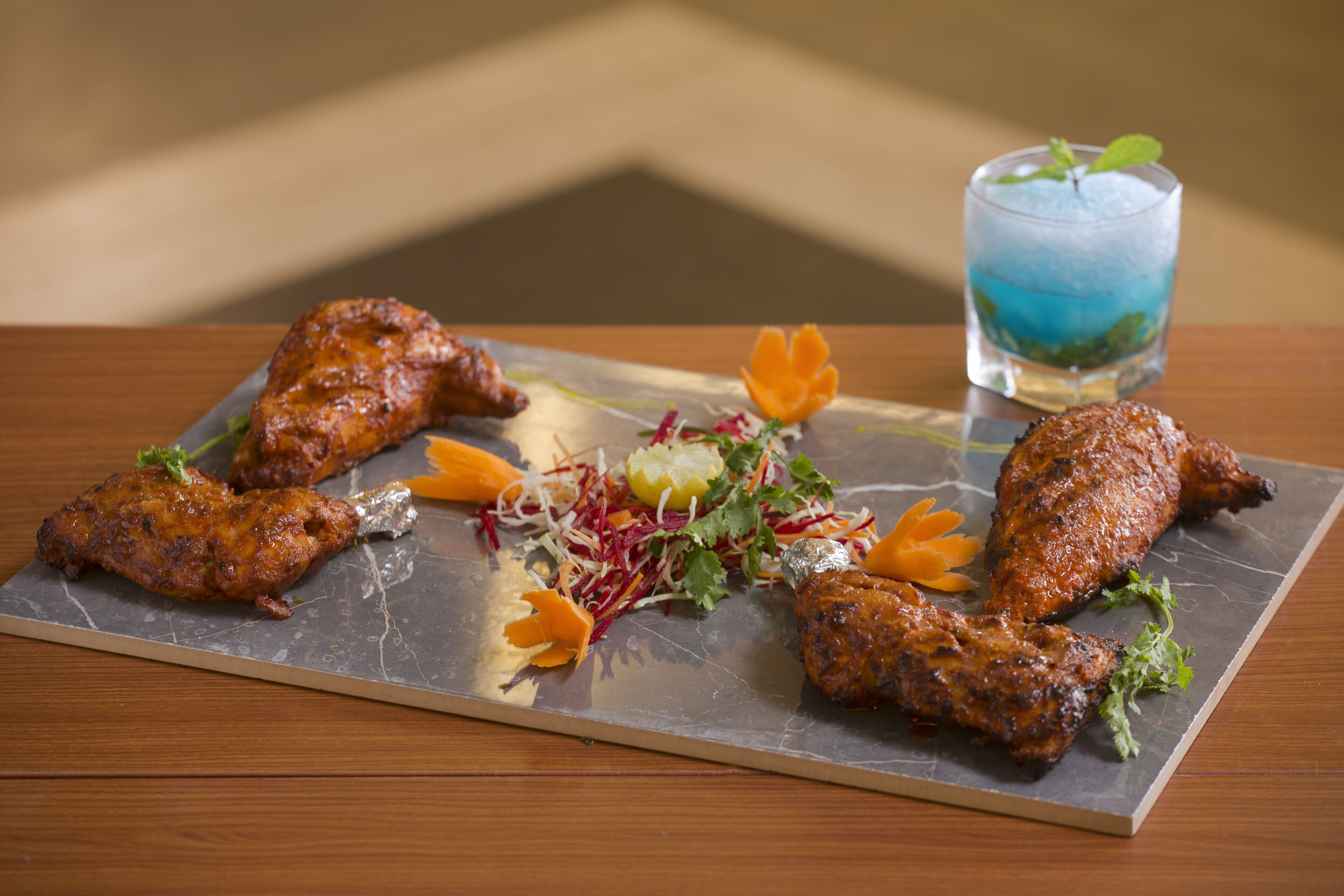 A dining table with a metal serving tray holding several pieces of grilled chicken, garnished with vegetables and a blue-colored drink.