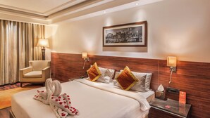 Quarto de hotel com uma cama bem arrumada, uma cadeira, um quadro com vista para a cidade e uma mesa de cabeceira com abajur.