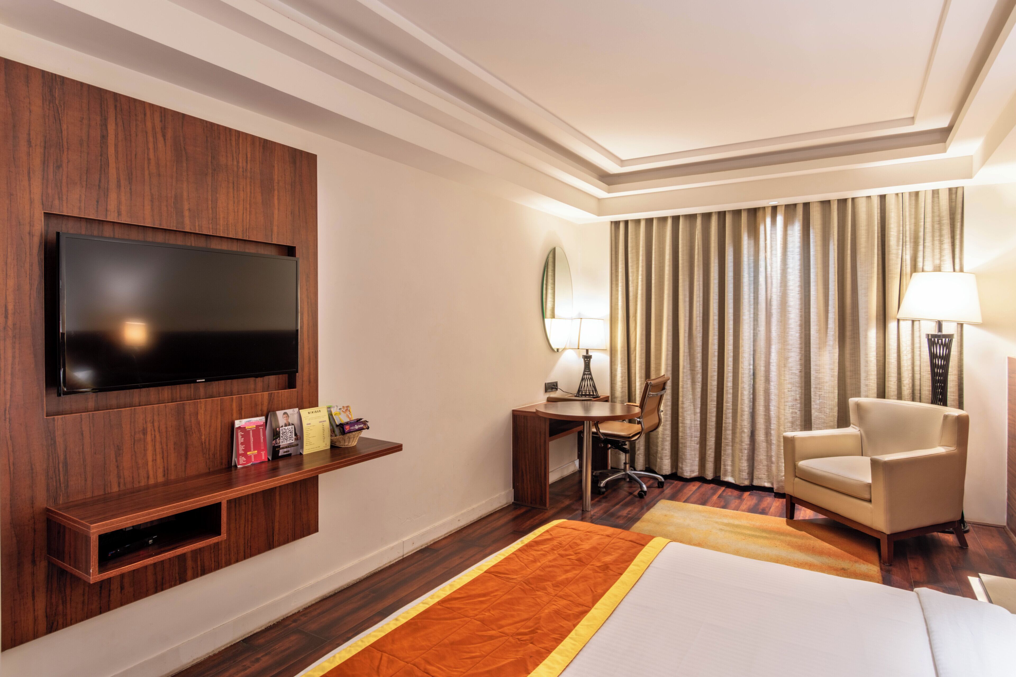 Habitación de hotel con televisor de pantalla plana montado en un panel de madera, un escritorio con una silla, una cama con cubrecama naranja y un ventanal con cortinas.