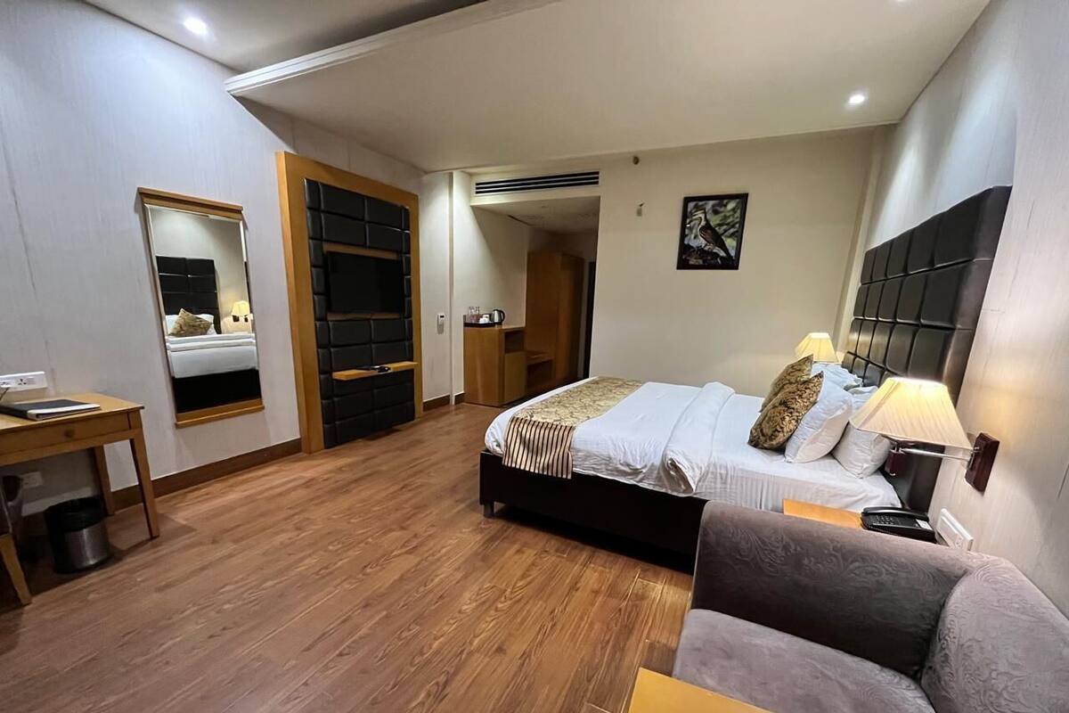 A modern hotel room with a wooden floor, a bed with a headboard, a sofa, a desk, and a mirror.