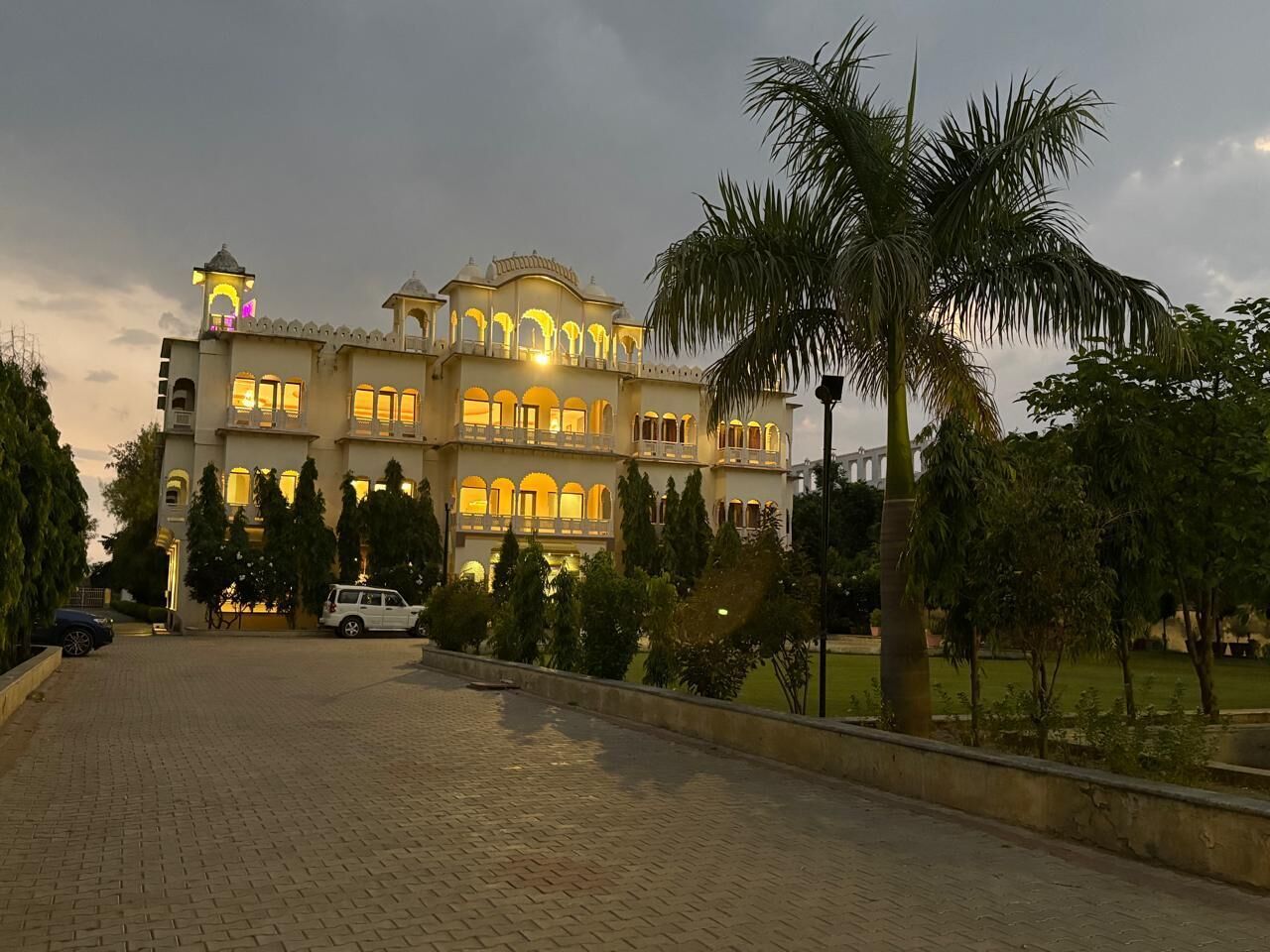 A large, multi-story building with arched windows and balconies, surrounded by greenery and palm trees.