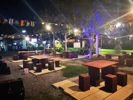 Un área de estar al aire libre con mesas y sillas, rodeada de follaje e iluminación con luces colgantes.