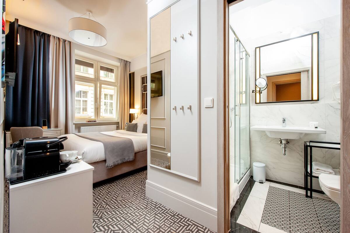 Habitación de hotel con cama, escritorio con cafetera, armario, baño con lavamanos y espejo, y alfombra estampada.