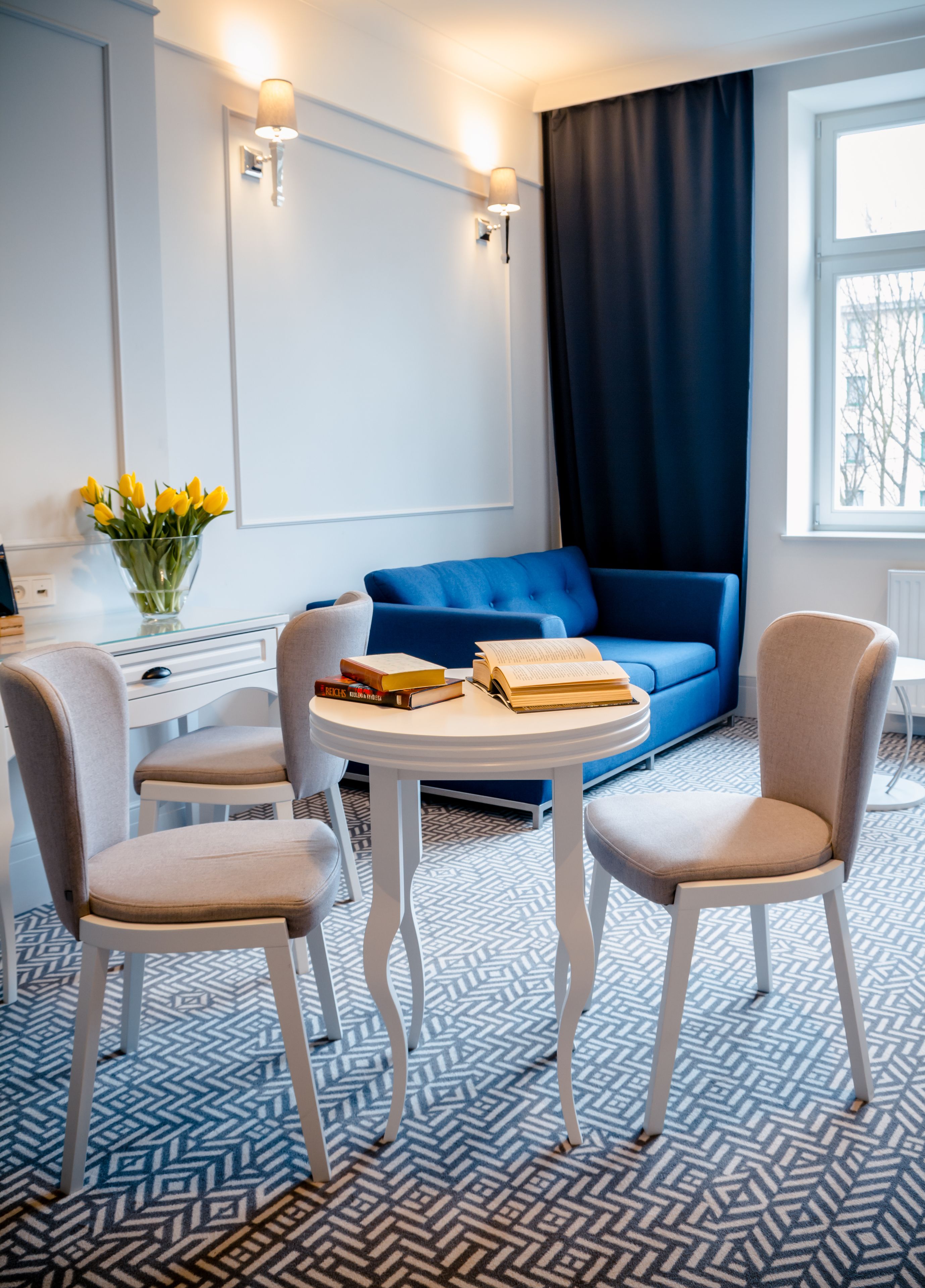 A modern room with a blue sofa, two white chairs, a round table with books, a vase with yellow flowers, and a window with a view of buildings.