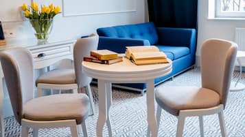 Ein modernes Zimmer mit einem blauen Sofa, zwei weißen Sesseln, einem runden Tisch mit Büchern, einer Vase mit gelben Blumen und einem Fenster mit Blick auf Gebäude.
