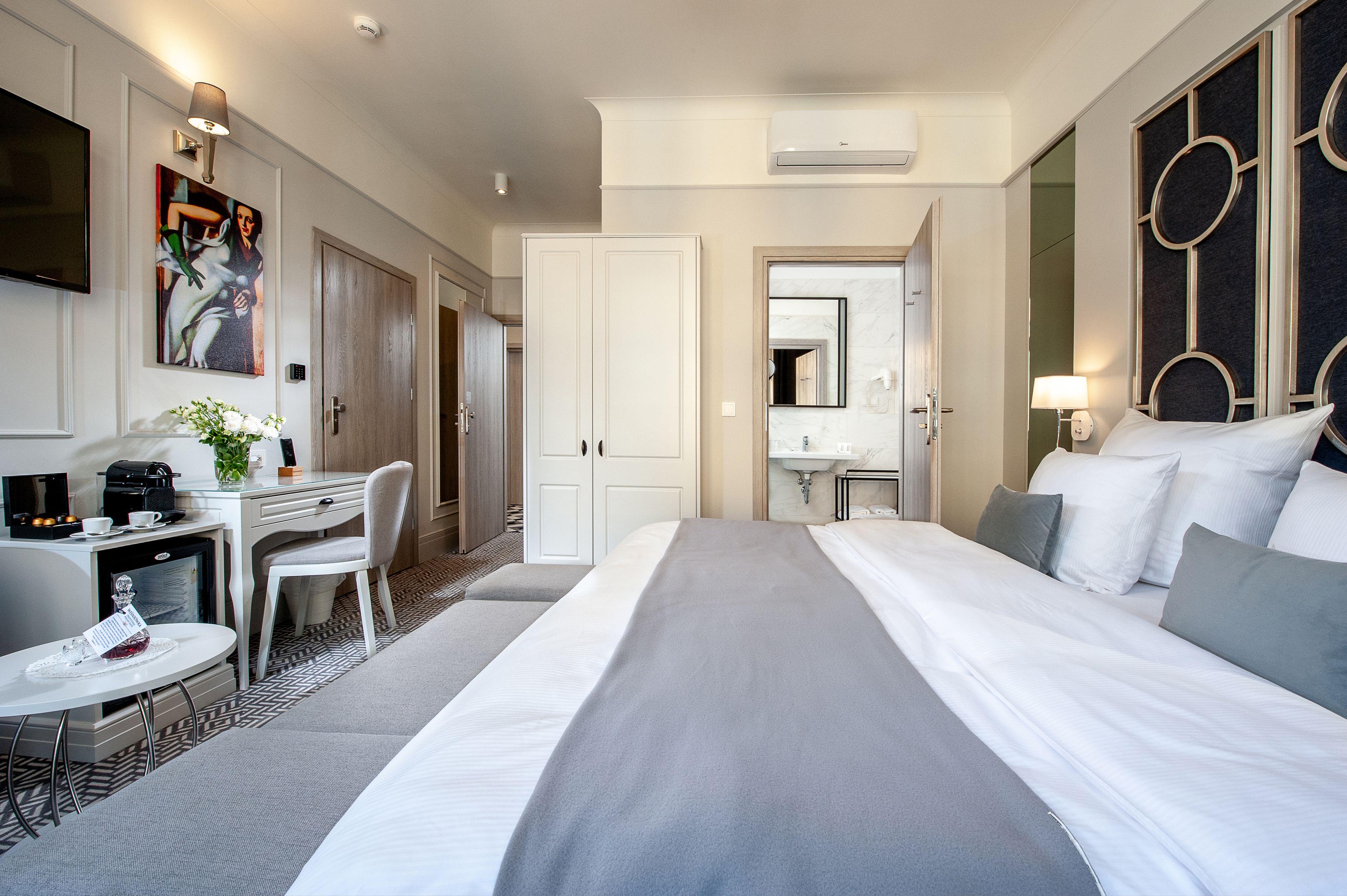 Habitación de hotel con cama, escritorio con silla, una mesita con bandeja, televisor y un cuadro en la pared.