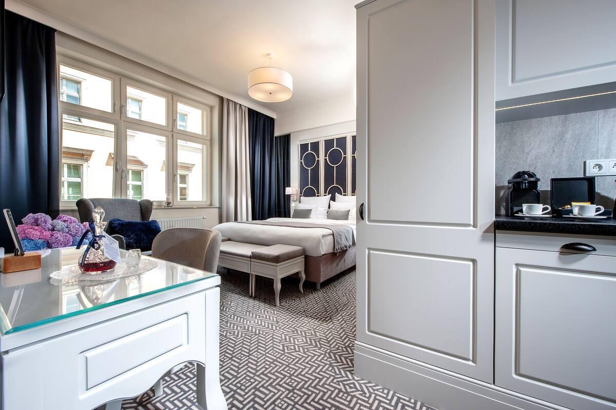 Habitación de hotel con cama, un escritorio con superficie de vidrio, una silla, un banco, una cafetera y vistas a edificios a través de ventanas amplias.