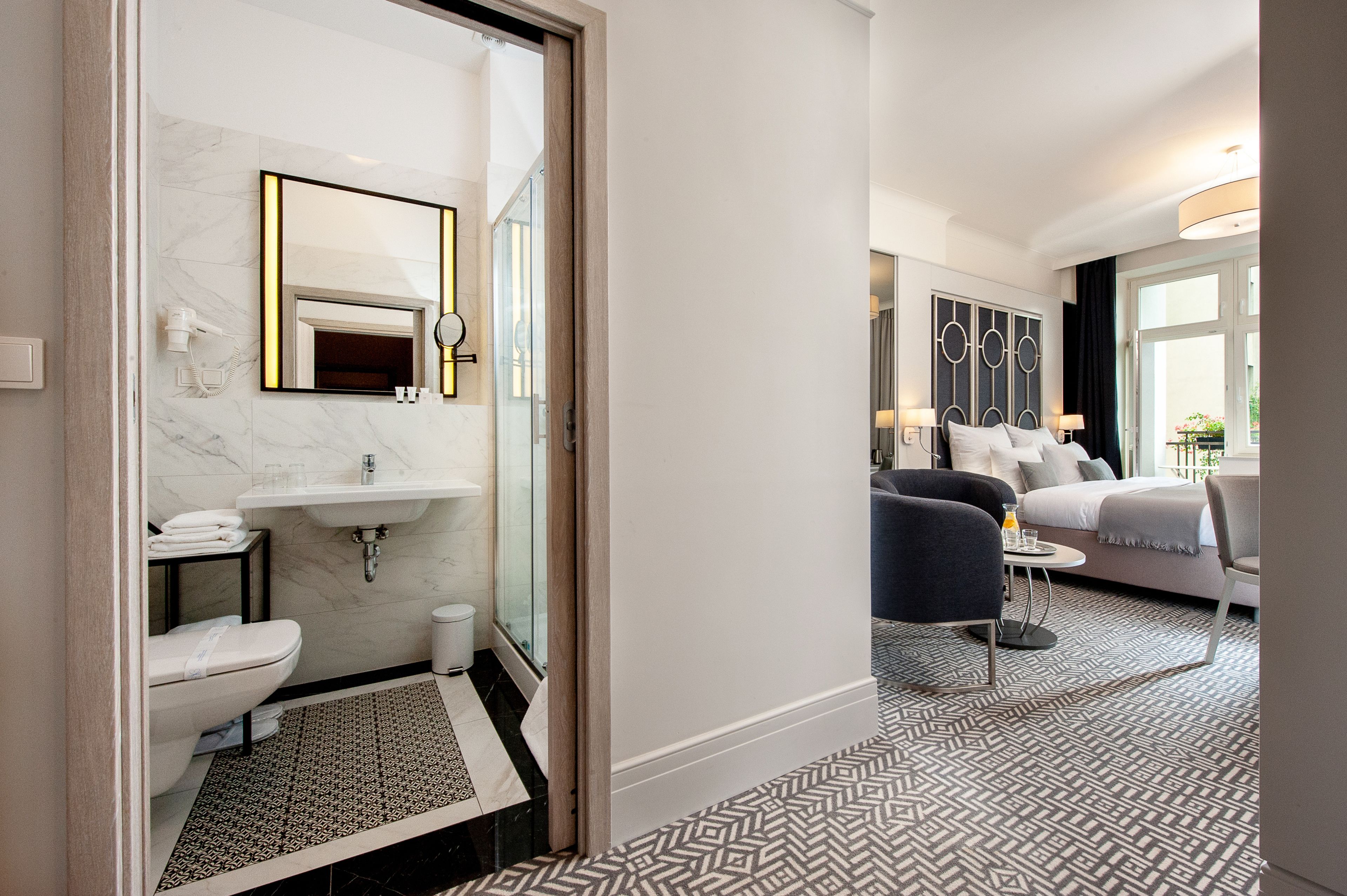 Une chambre d’hôtel moderne dotée d’une salle de bain avec un lavabo en marbre, des toilettes et une douche. La chambre comprend un grand lit, une table de chevet et offre une vue sur l’extérieur grâce à une fenêtre.