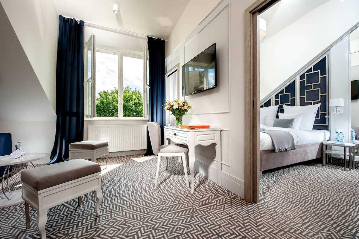 Habitación de hotel con cama, escritorio, silla, banca, televisor y ventana con cortinas.