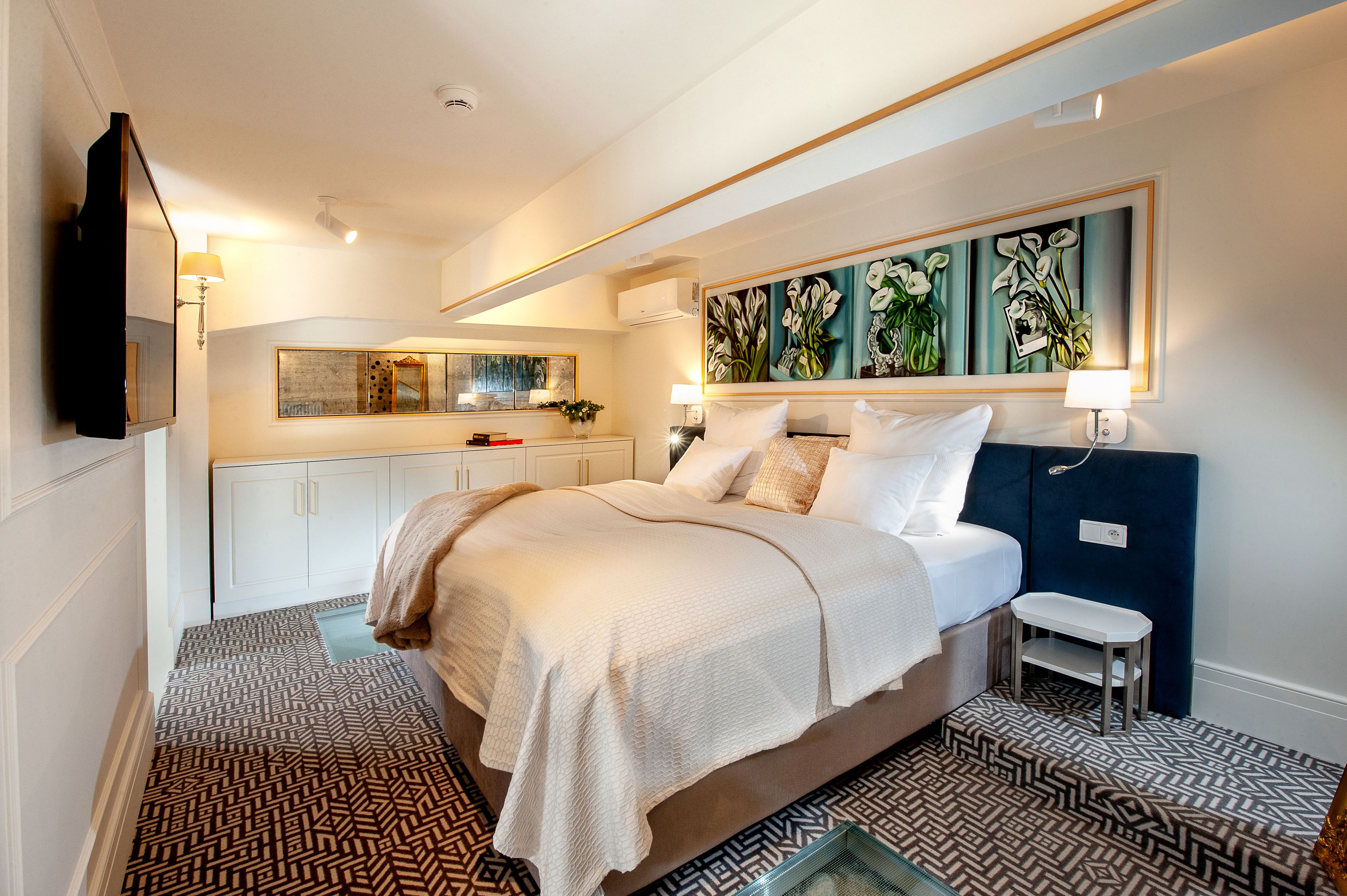 Una habitación de hotel moderna con una cama grande, una mesita de noche pequeña, un espejo y un cuadro sobre la cama.