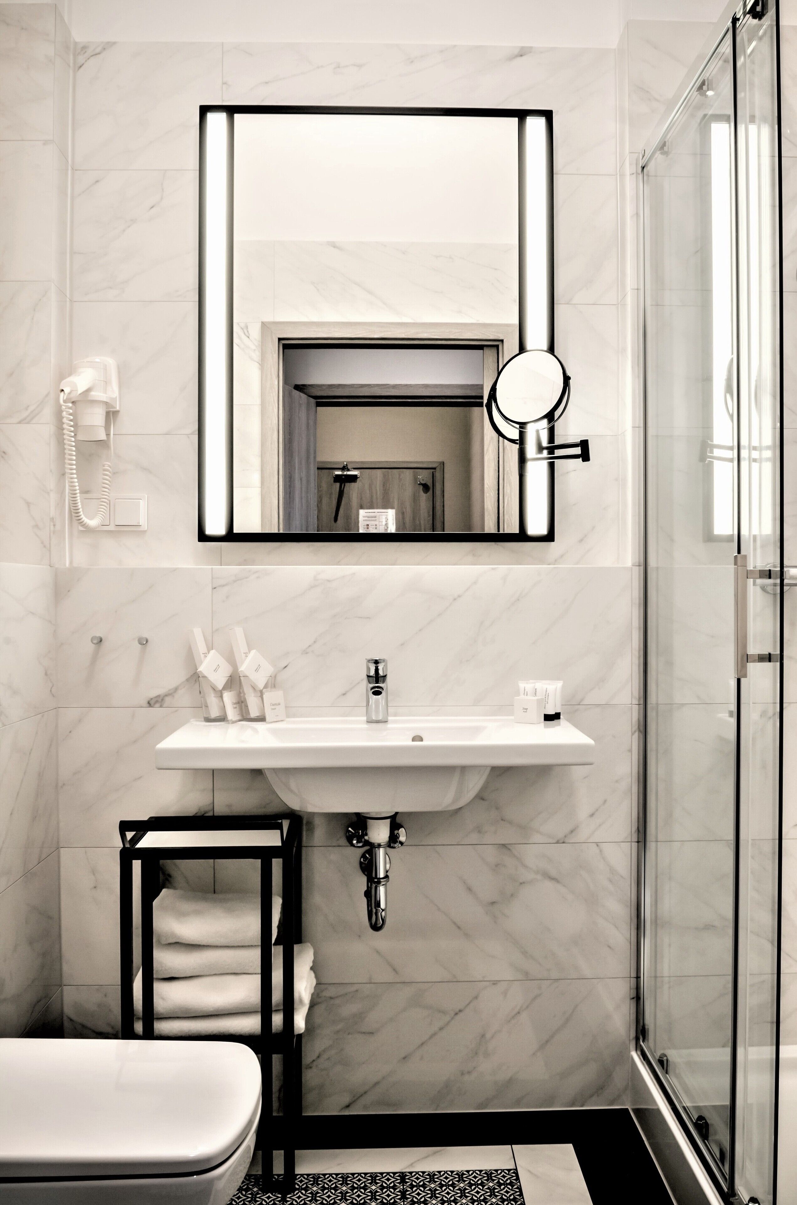 A modern bathroom with a marble wall, a white sink, a black towel rack, and a glass shower enclosure.
