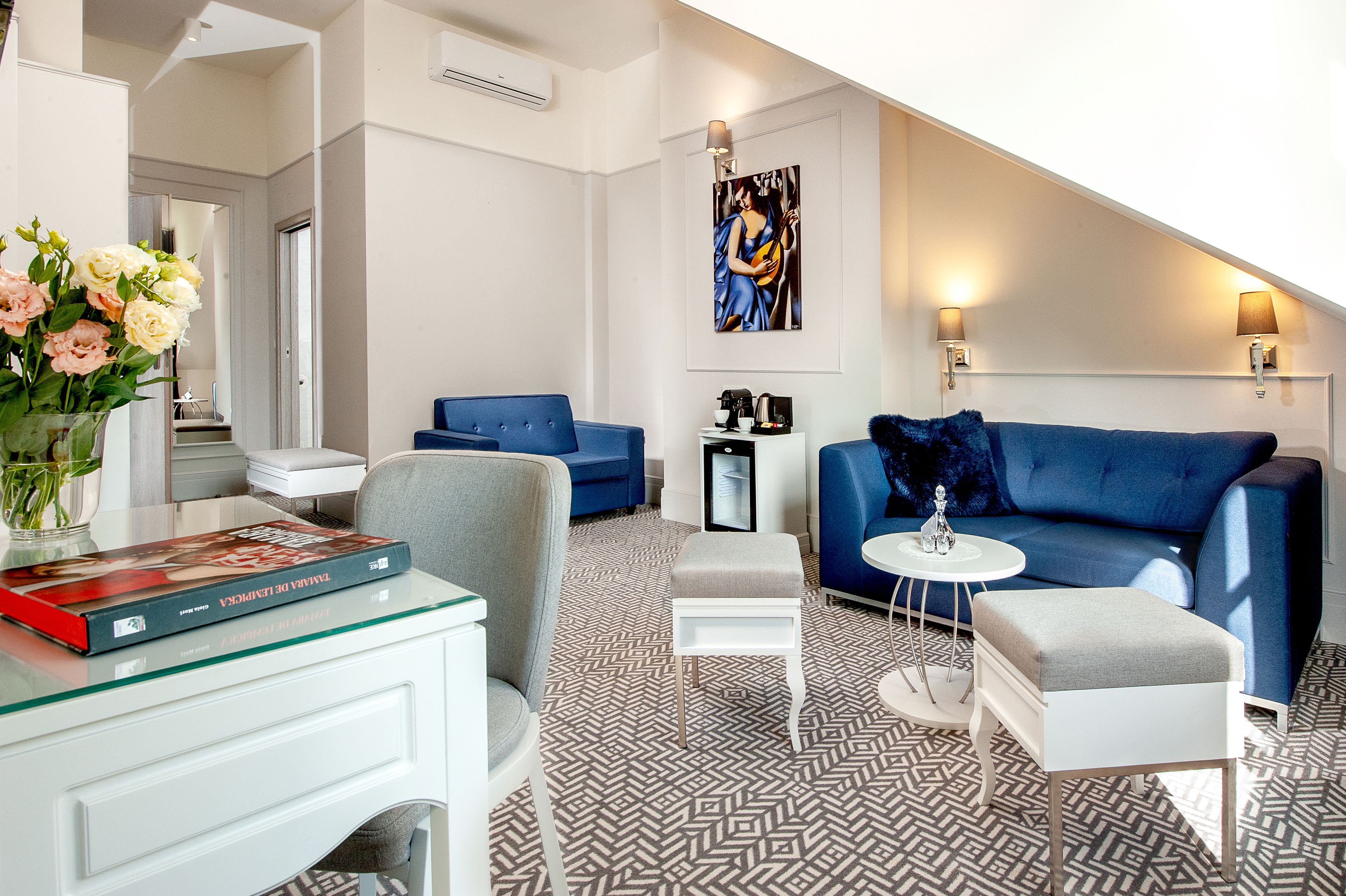 A modern living room with a blue sofa, a small white table, and a grey armchair. A book and a vase of flowers are on a glass table.