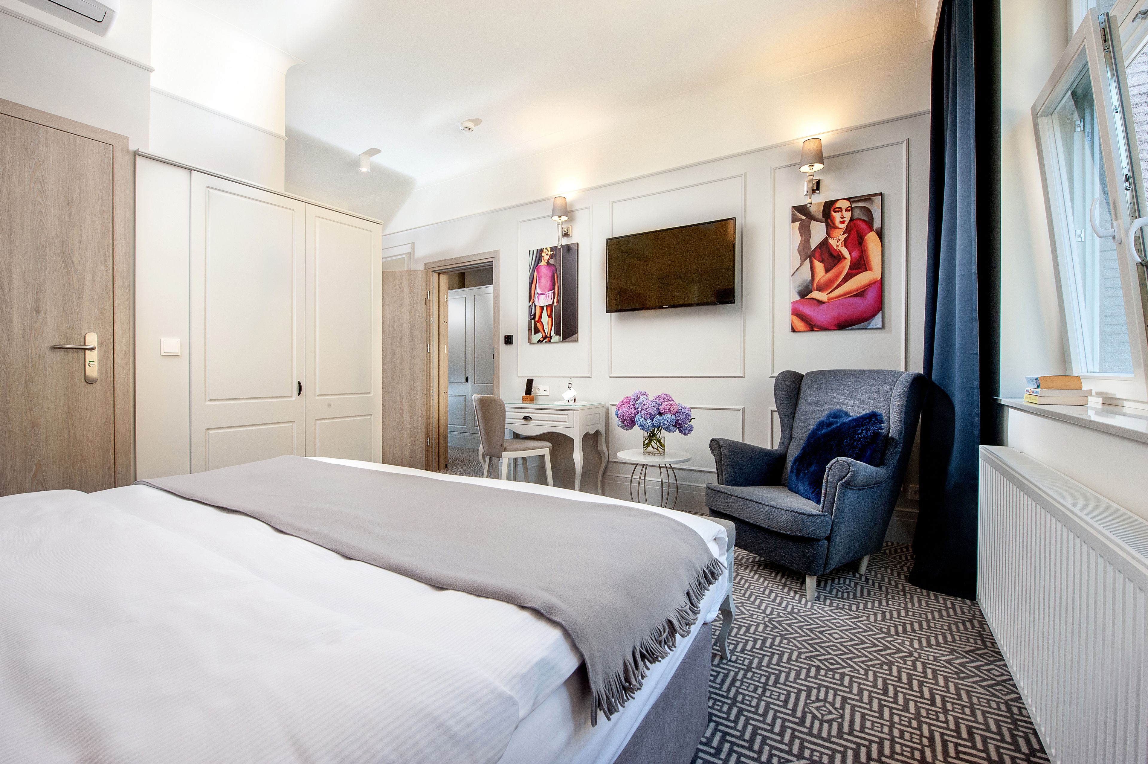 A hotel room with a bed, a desk, a chair, a TV, and artwork on the wall.