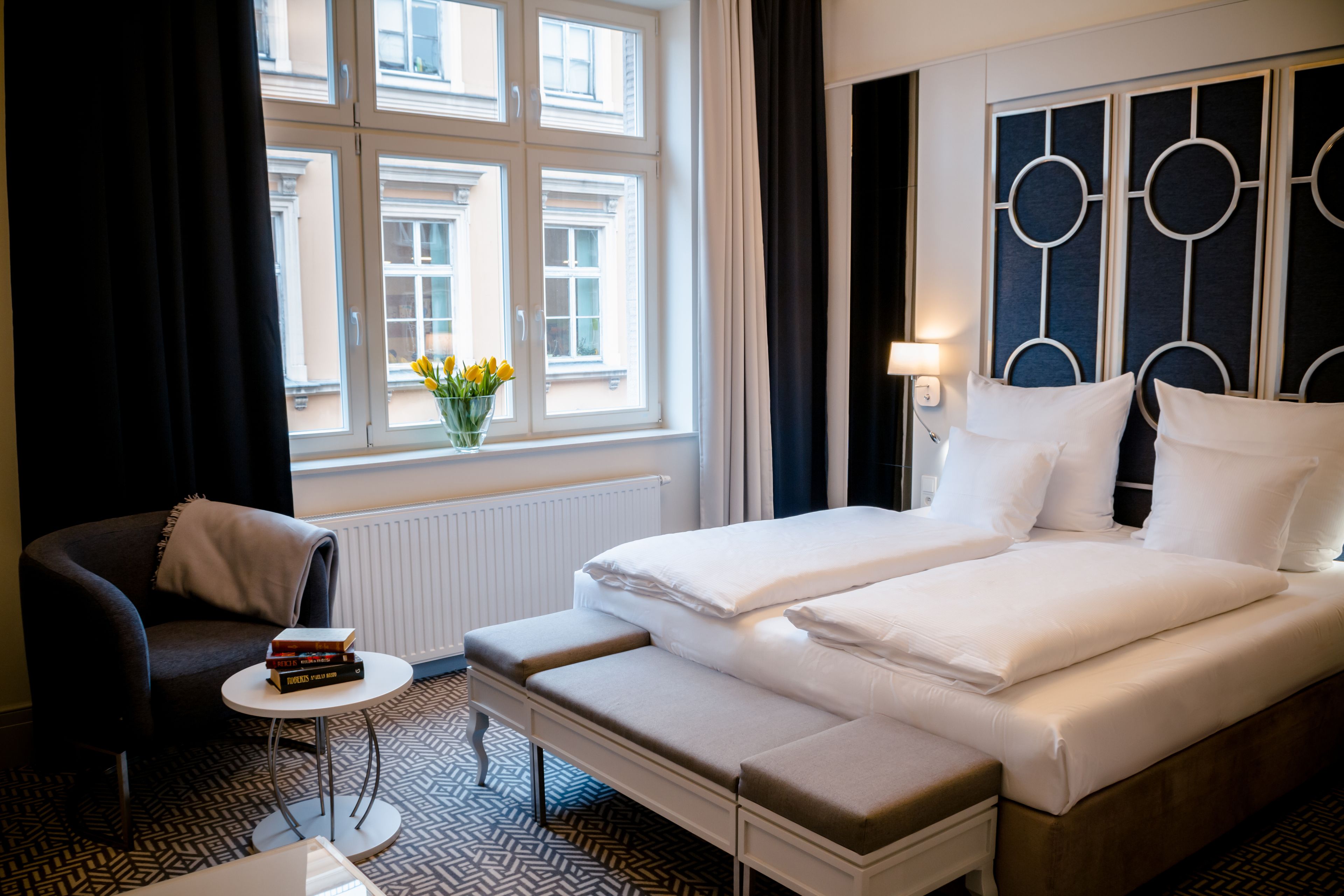 A hotel room with a bed, bedside table, chair, and a window with curtains.