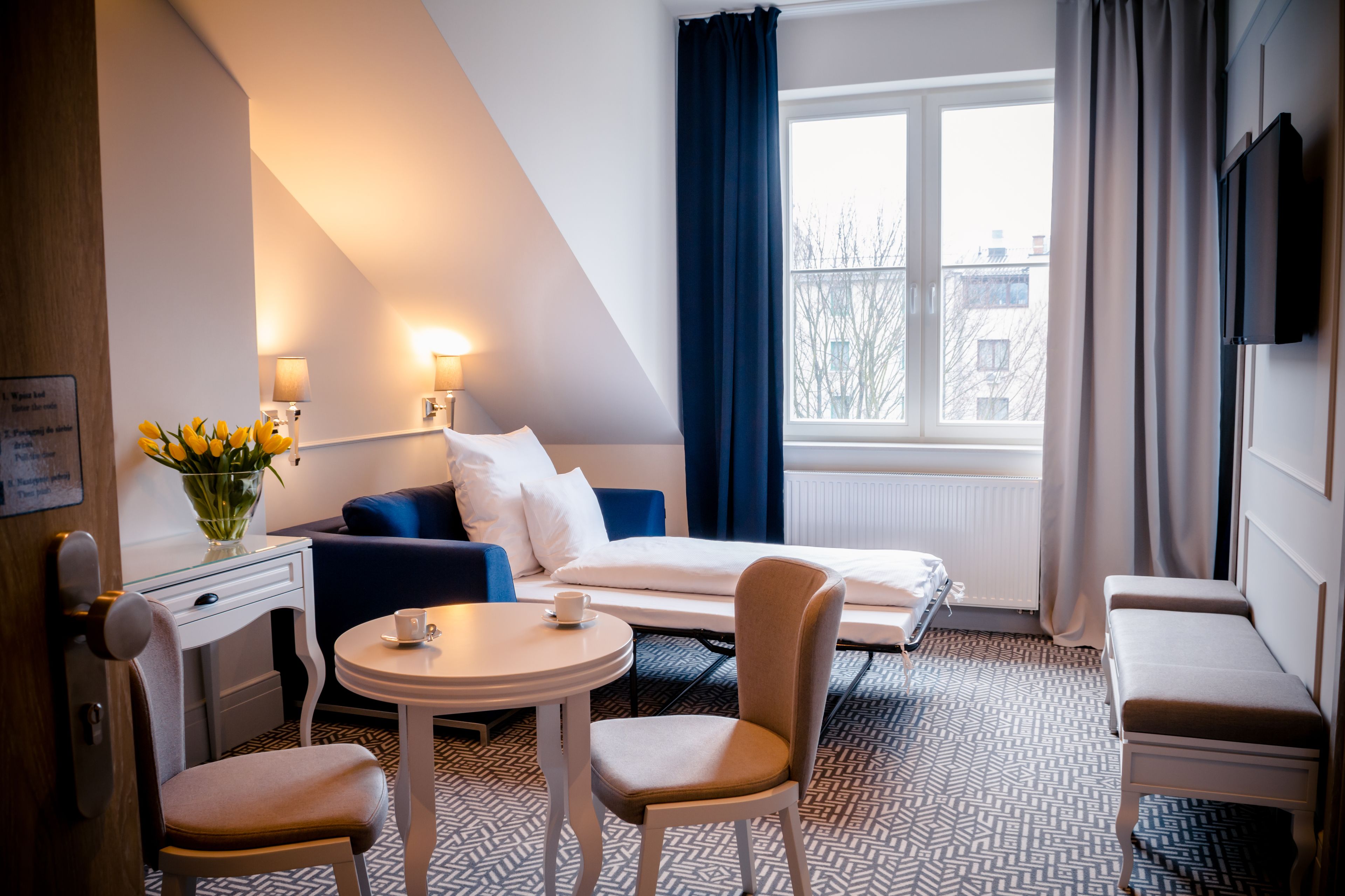 A hotel room with a bed, a sofa, a dining table, and a chair. There is a window with curtains and a view of buildings outside.