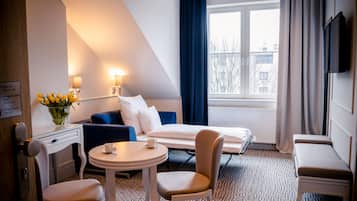 A hotel room with a bed, a sofa, a dining table, and a chair. There is a window with curtains and a view of buildings outside.
