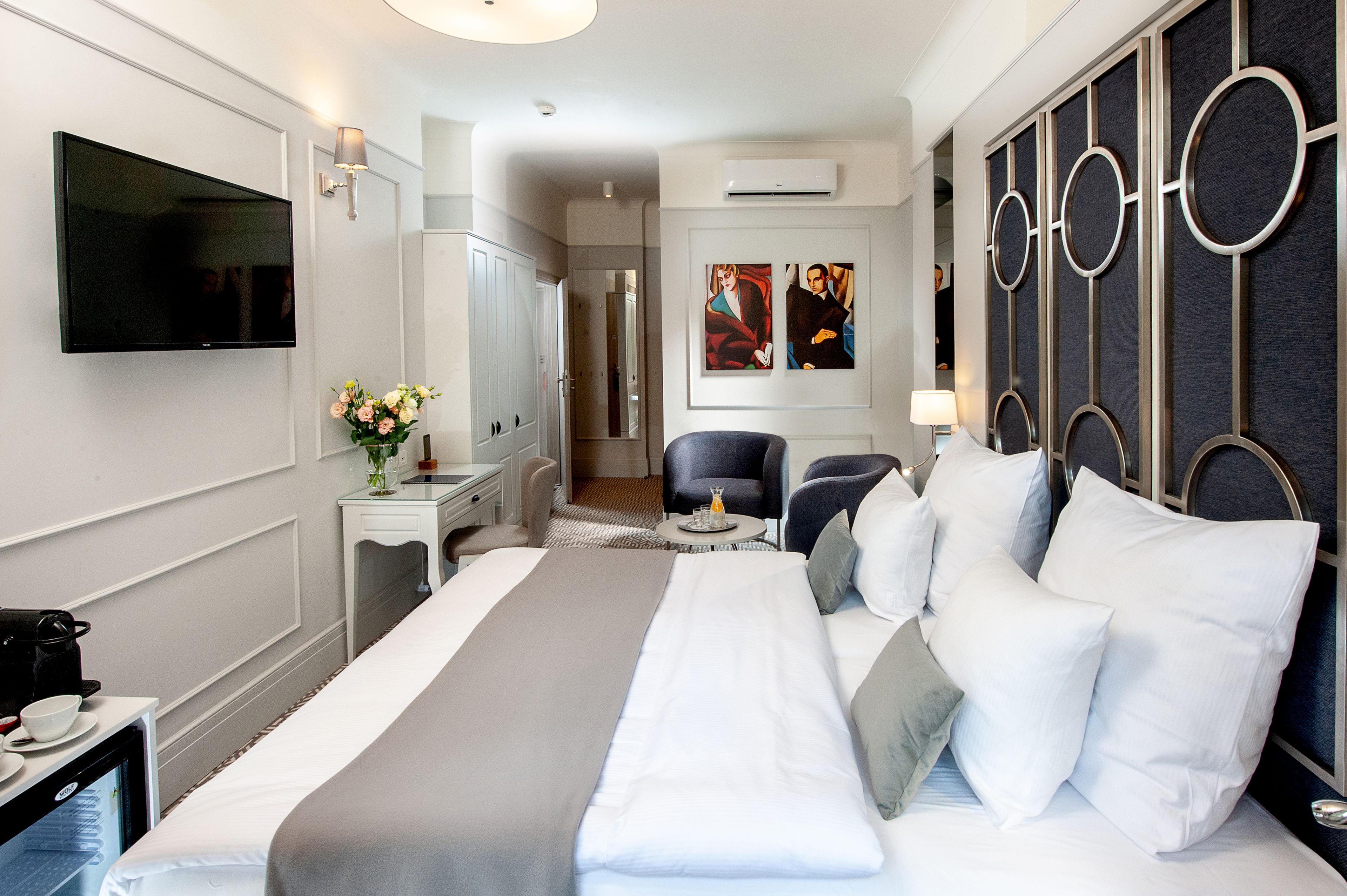 A hotel room with a large bed, a television, a desk with a lamp, a chair, and artwork on the wall.