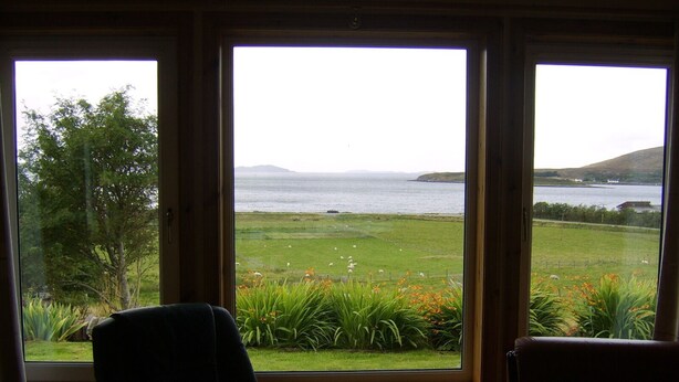 A room with a large window overlooking a grassy field and a body of water. - Top House (Ullapool)