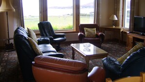A cozy living room with a sofa, armchairs, a coffee table, and a television. Large windows offer a view of a field and mountains. - Top House (Ullapool)