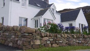 A white two-story house with a stone wall in front, a pitched roof, and a stone chimney. - Top House (Ullapool)