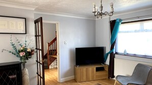 A living room with a TV, wooden furniture, a staircase, and a window with curtains. - Huku Kwetu - Chelsea Garden (Dunstable)