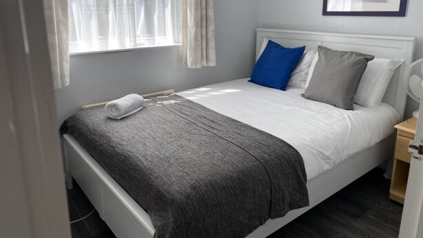 A neatly made bed with a white comforter and gray throw blanket, a blue pillow, and a gray pillow. There is a framed picture on the wall above the bed. - Huku Kwetu - Chelsea Garden (Dunstable)