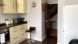 A compact kitchen with a toaster, microwave, and oven. A bar stool is positioned near the counter. A clock with a coffee cup design hangs on the wall. - Huku Kwetu - Chelsea Garden (Dunstable)