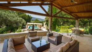 An outdoor patio with wicker furniture, a glass coffee table, and a view of a pool and garden. - Domaine Les Roullets (Oppede)