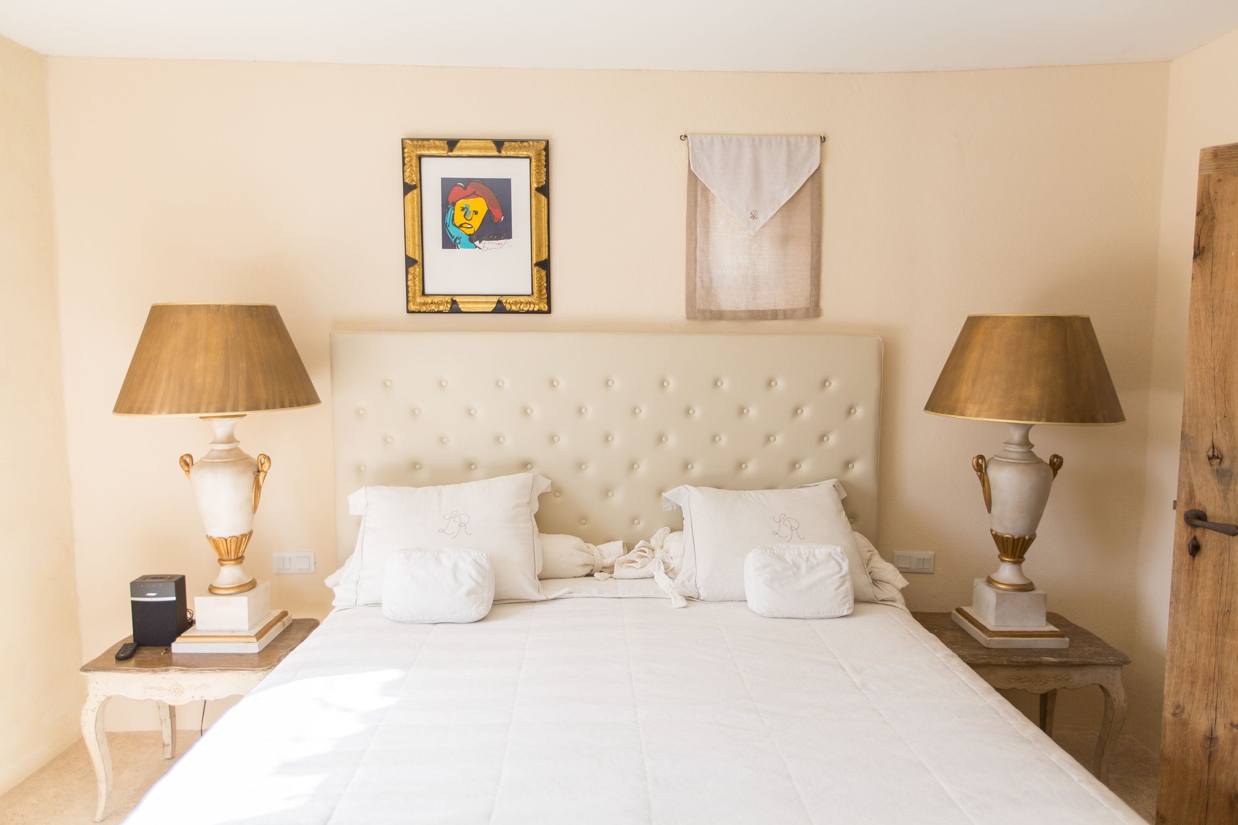 A neatly arranged bedroom with a large bed, two bedside tables with lamps, a framed picture on the wall, and a door on the right.