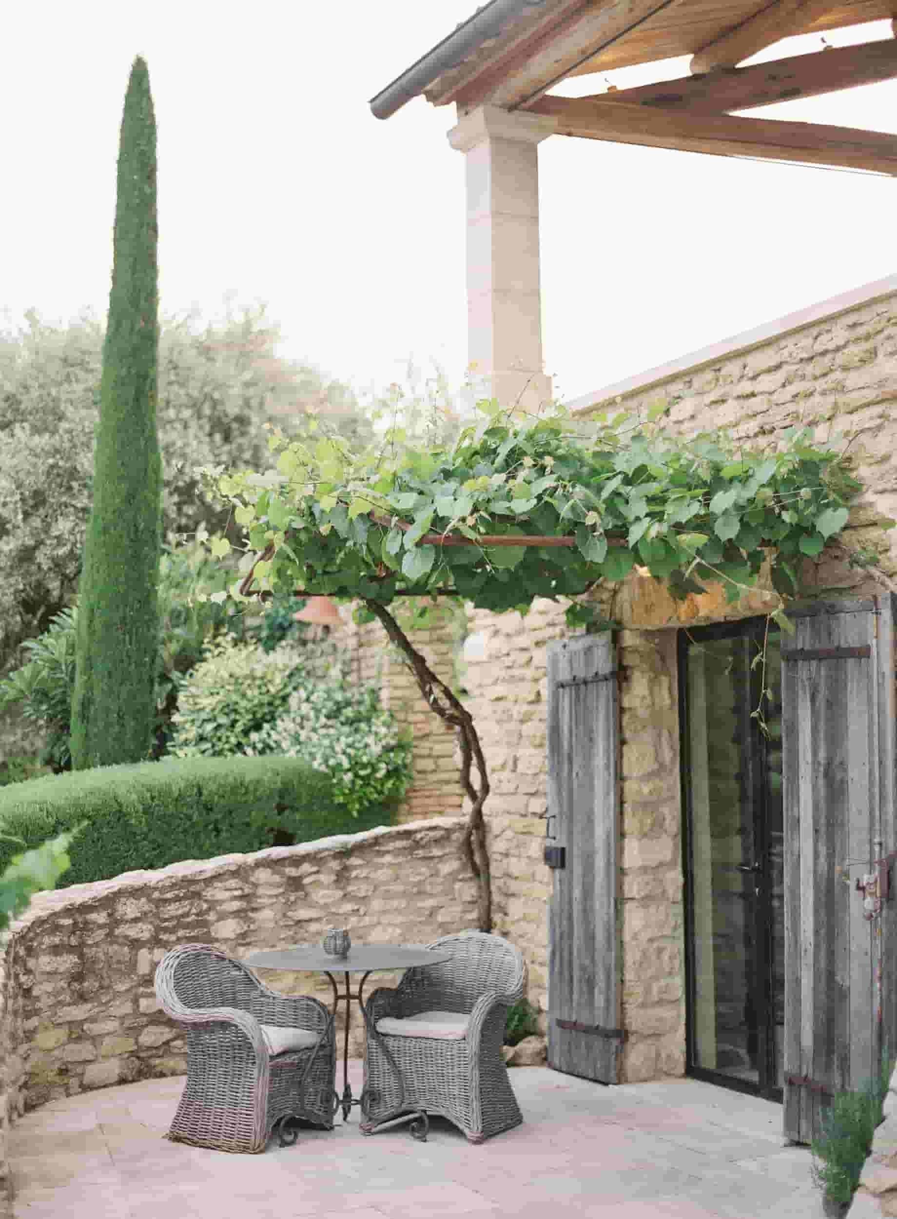 Une terrasse extérieure avec des meubles en osier, une pergola recouverte de plantes grimpantes et un mur en pierre.