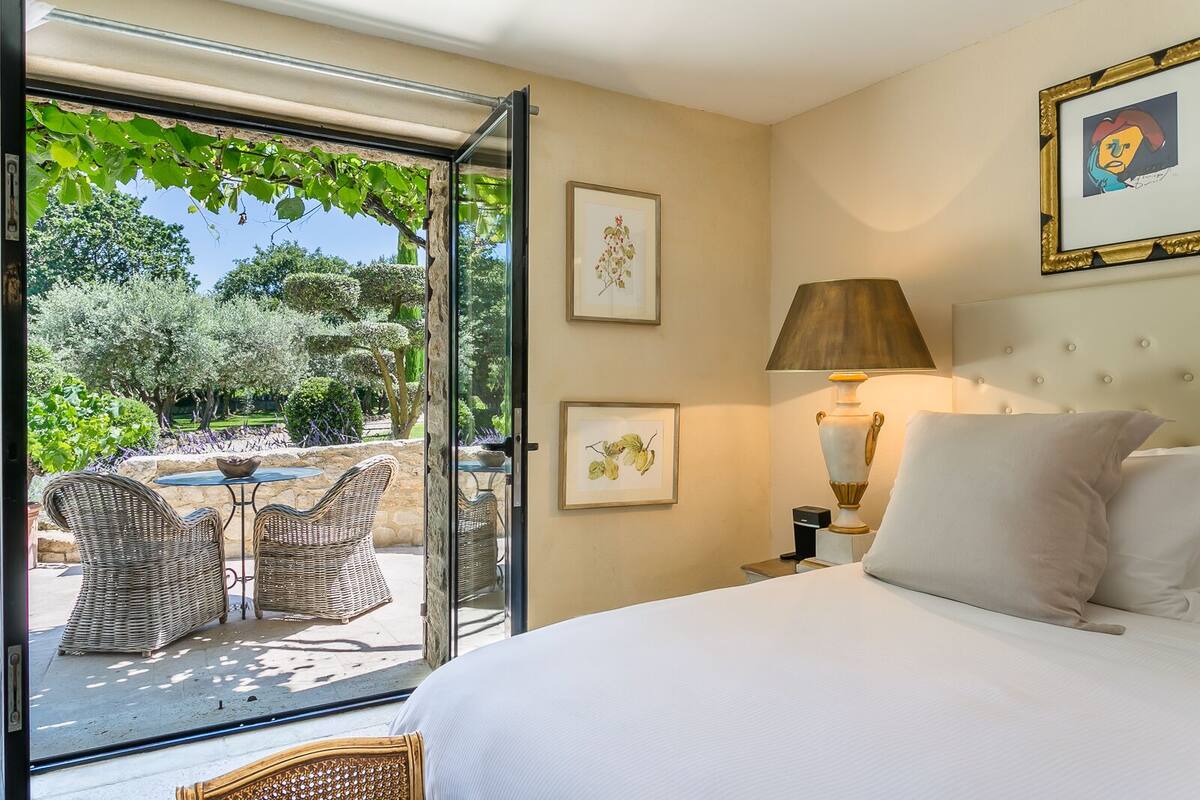 Quarto com cama, mesa de cabeceira, luminária e obras de arte emolduradas. Há uma porta de vidro que dá para uma área externa com mobiliário de vime.