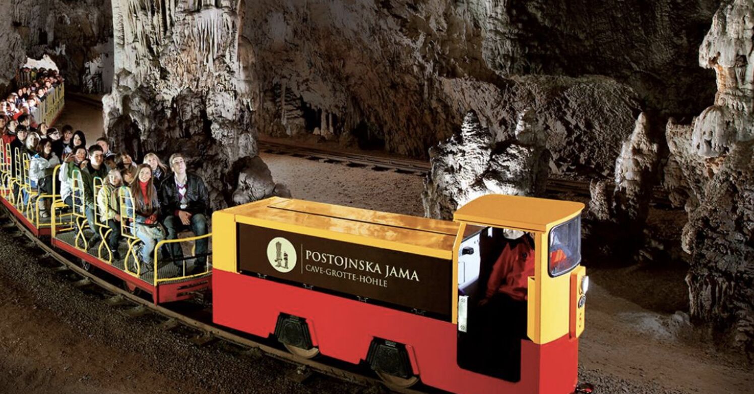 A tourist train with passengers, labeled "POSTOJNSKA JAMA CAVE-GROTTEN-HÖHLE," traveling through a cave.