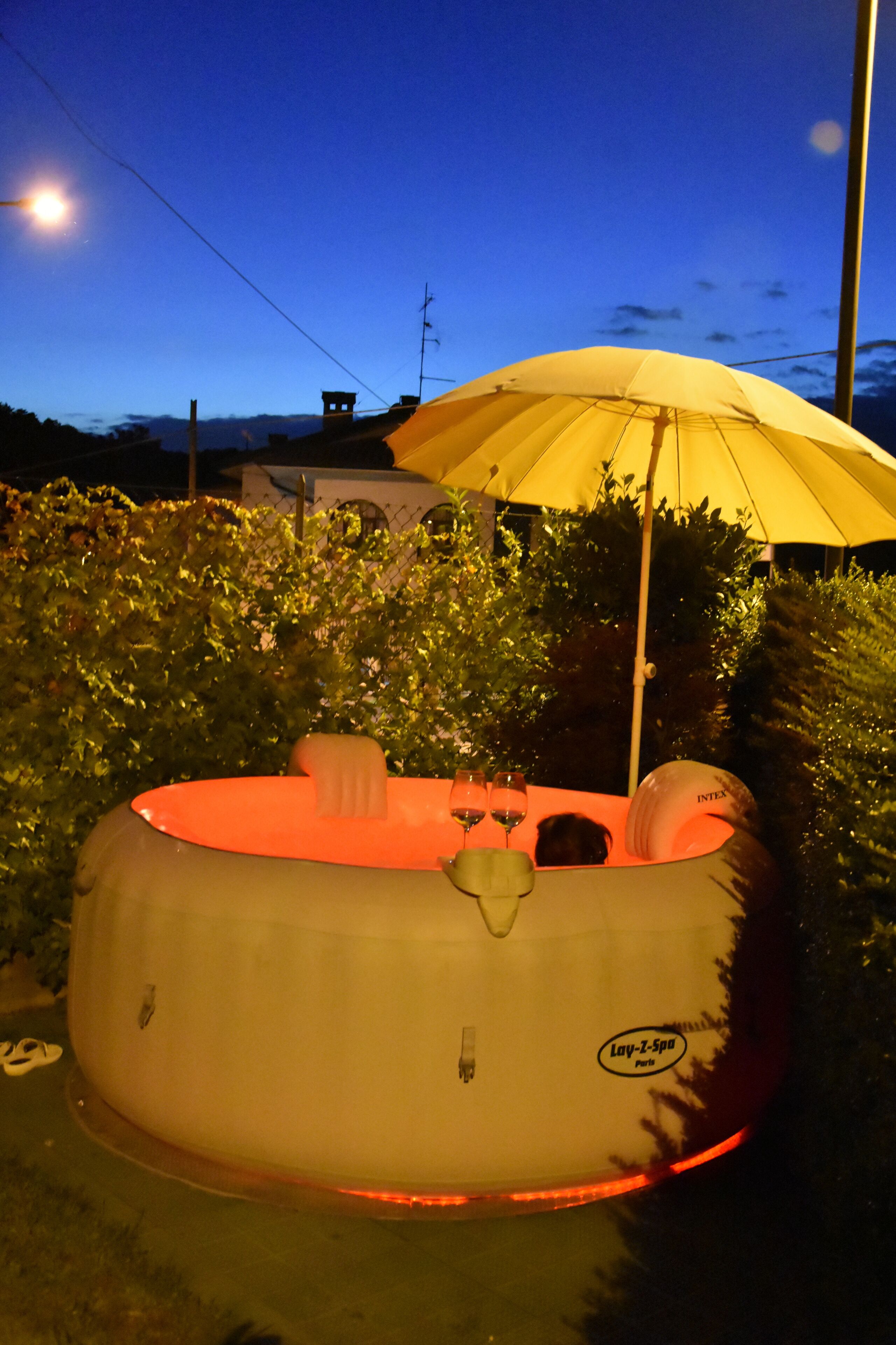 An inflatable hot tub with a yellow umbrella, surrounded by greenery.