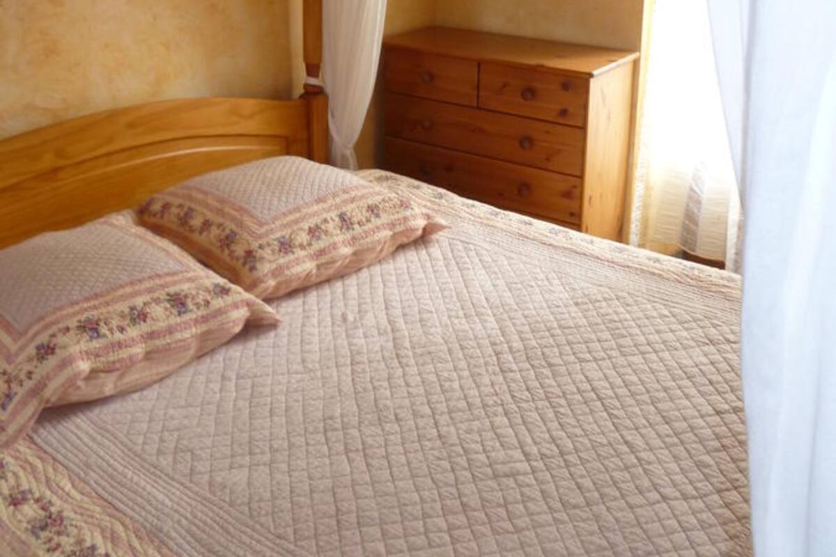 A bedroom with a wooden bed frame, a bedside table, a mirror, and a window with curtains.