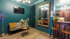 A teal-walled room with a sofa, a TV, a large mirror, and a bar area with stools. - HOTEL MAAT ETOILE (Paris)
