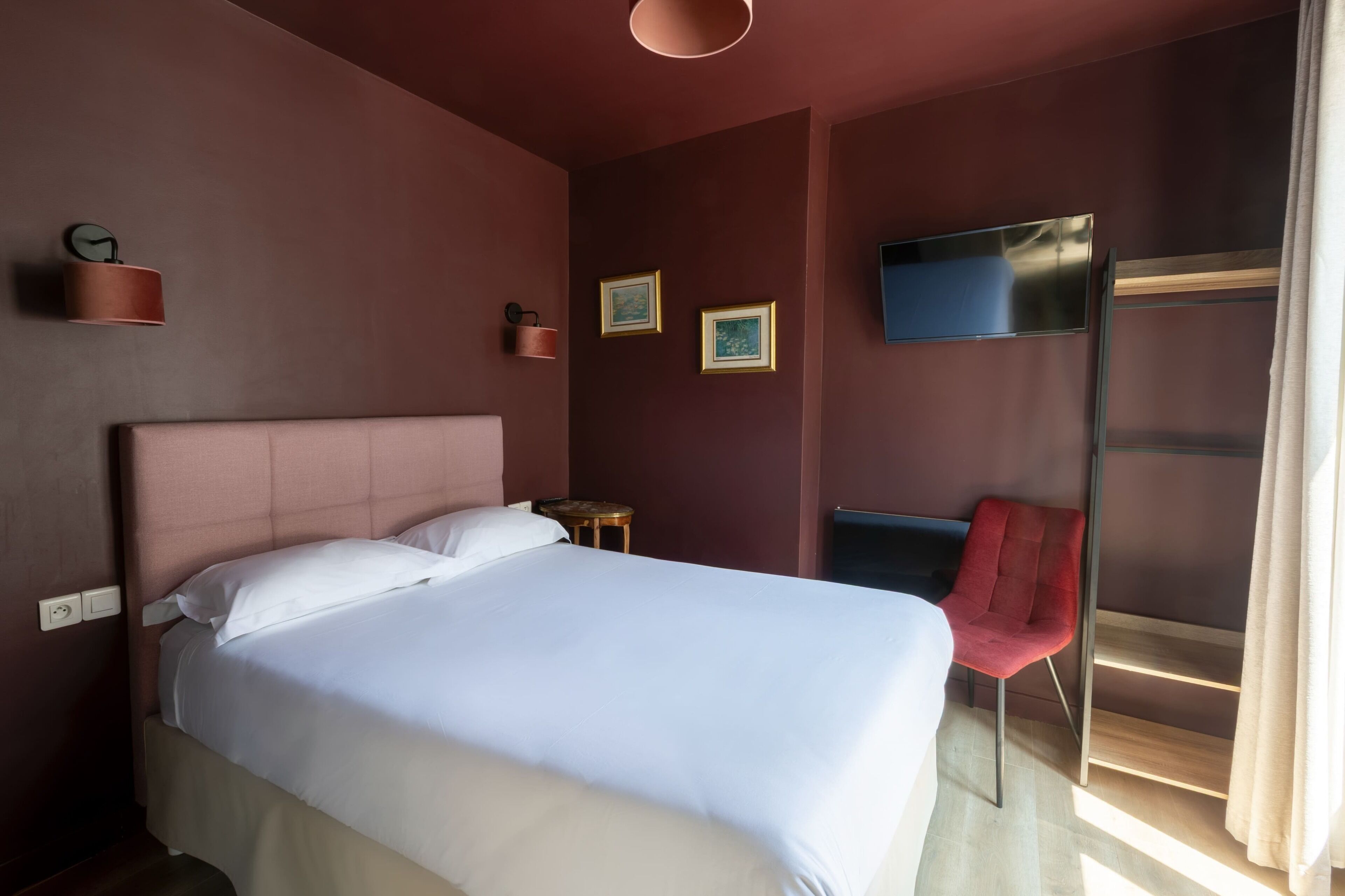 A hotel room with a large bed, a red chair, a TV mounted on the wall, and a small table.