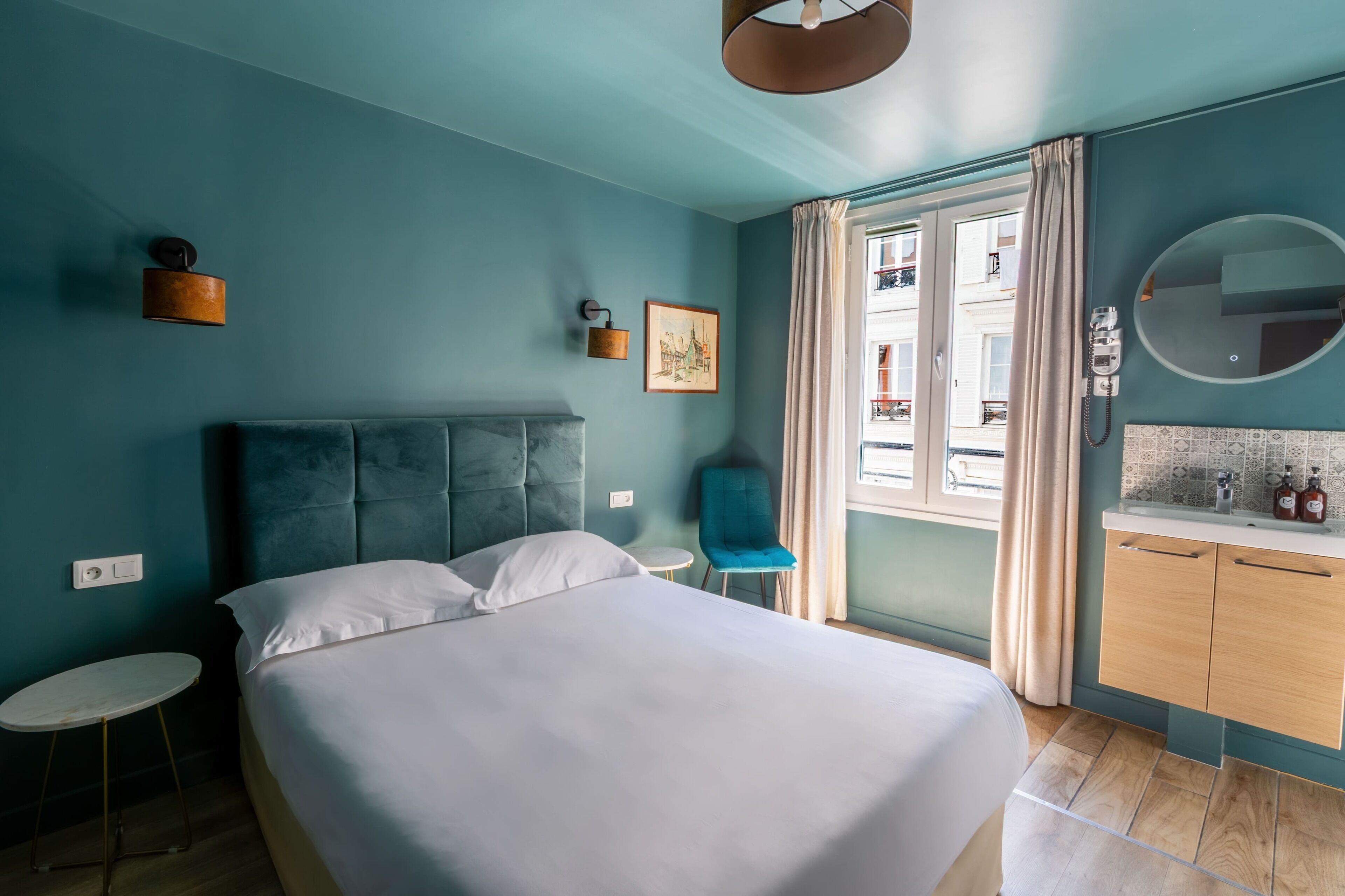 Un dormitorio con cama, un espejo redondo, una mesita pequeña y redonda, una silla azul y vistas al exterior a través de una ventana.