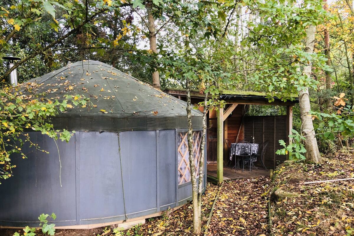 Een joert met een overdekt dak, omringd door bomen en een houten constructie met een tafel en stoelen.