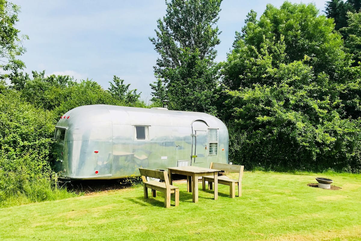 Een zilveren caravan met een klein raam en een deur, omringd door weelderige begroeiing en een picknicktafel met bankjes.