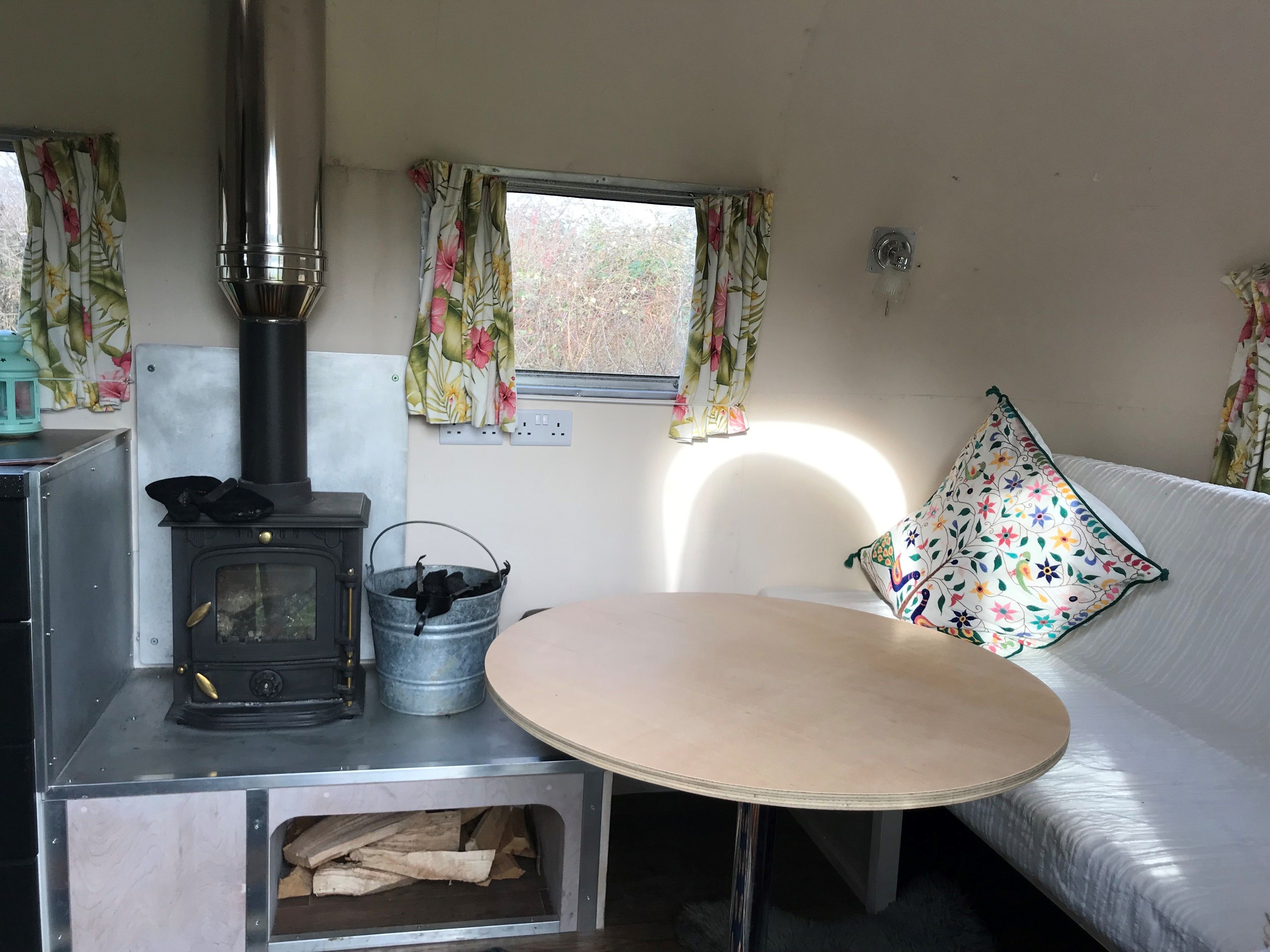 Un intérieur chaleureux avec un poêle à bois, un seau en métal, un canapé et une fenêtre ornée de rideaux à motifs floraux.