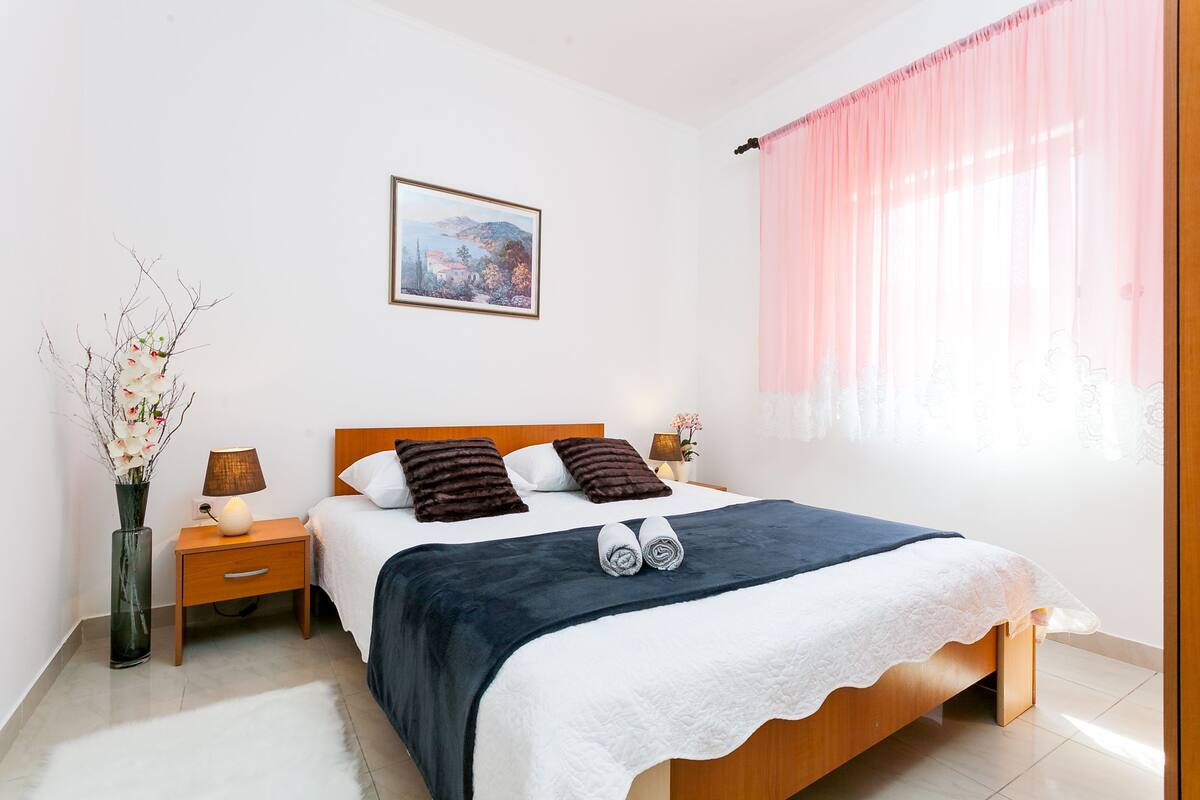 Quarto bem arrumado com cama, mesas de cabeceira, abajur, um vaso com flores e um quadro na parede.