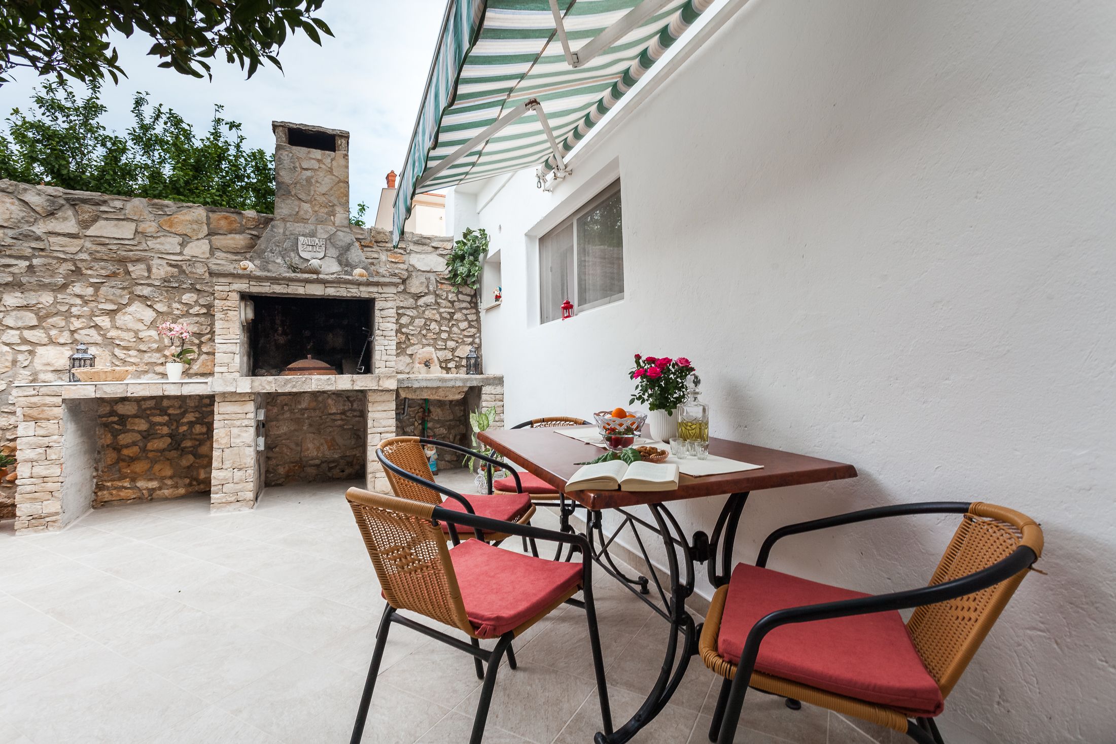 An outdoor patio with stone walls, a brick oven, wicker chairs, and a table with a book and flowers.