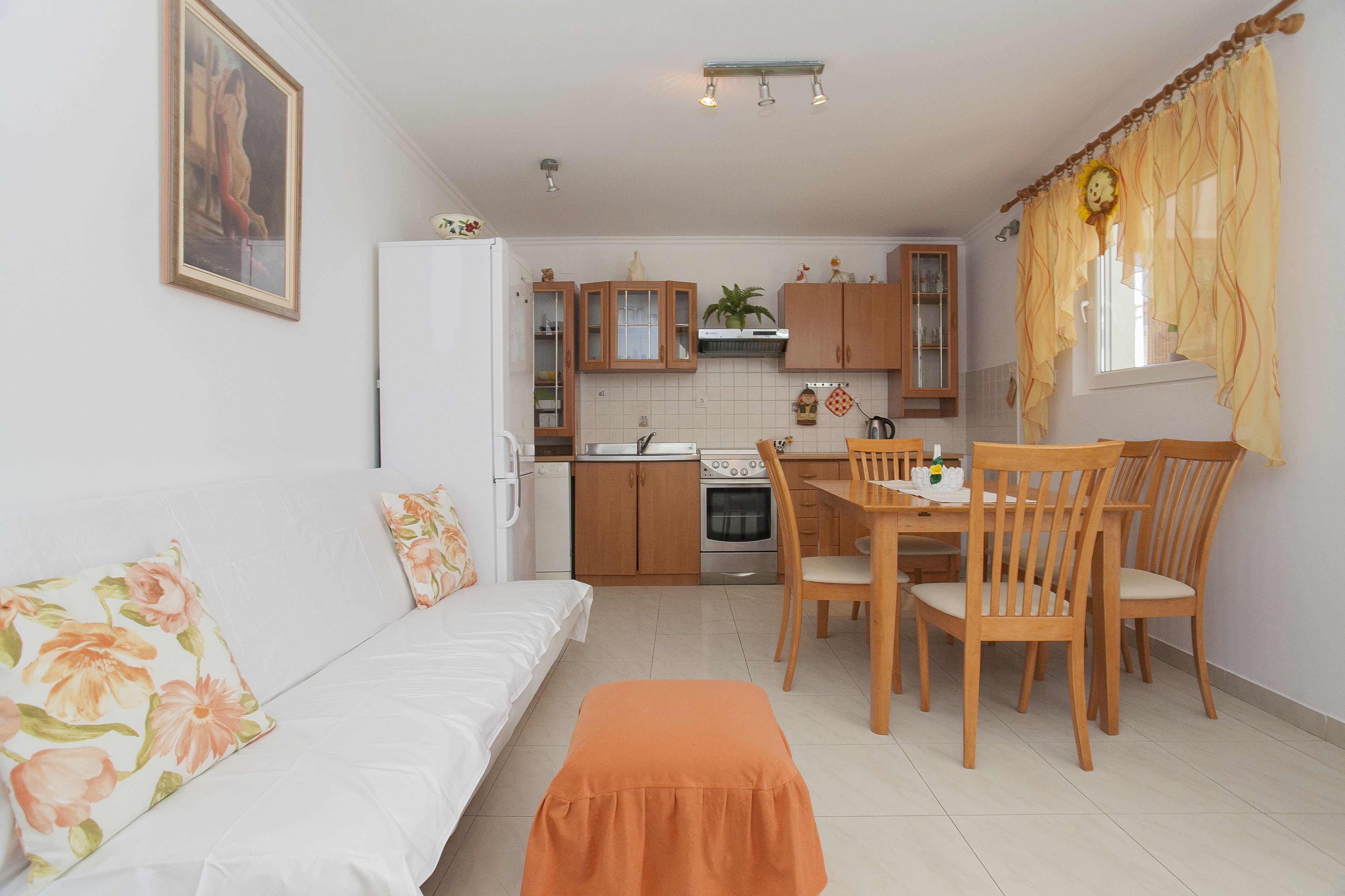 Une cuisine compacte avec des meubles en bois, une table à manger avec des chaises, un canapé blanc agrémenté de coussins à motifs floraux et un petit banc orange.