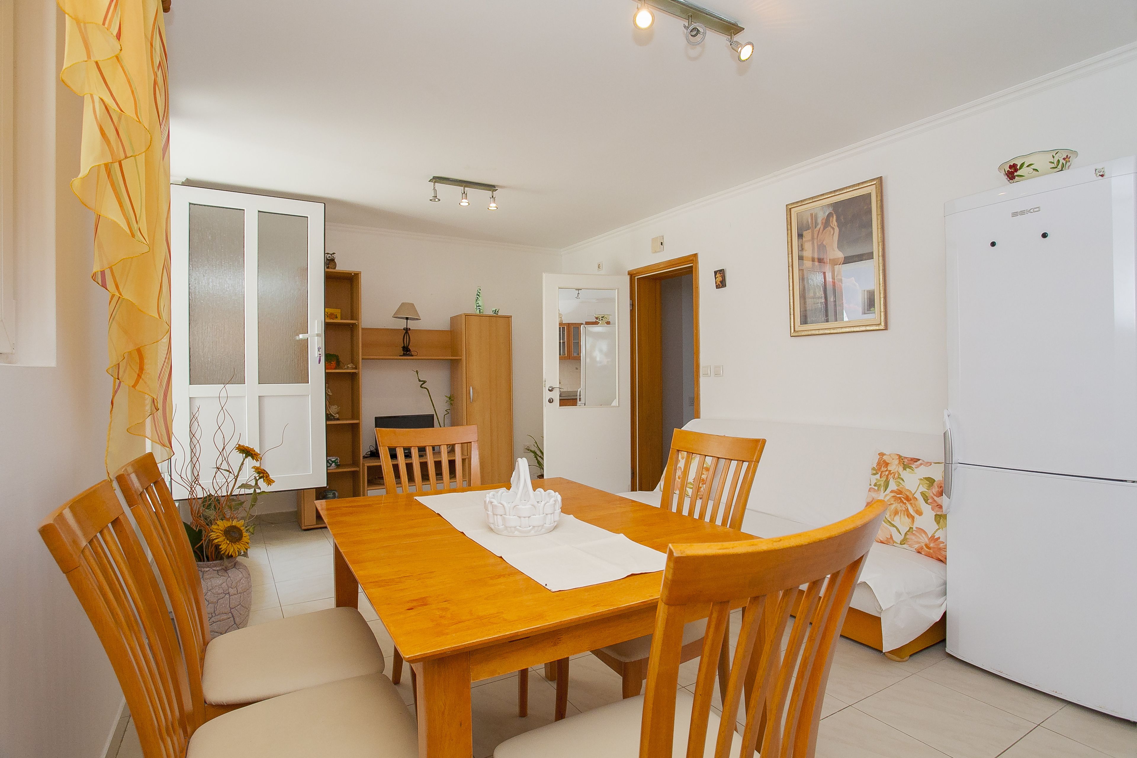 Uma área de refeições com uma mesa e cadeiras de madeira, uma geladeira branca e uma cozinha ao fundo.