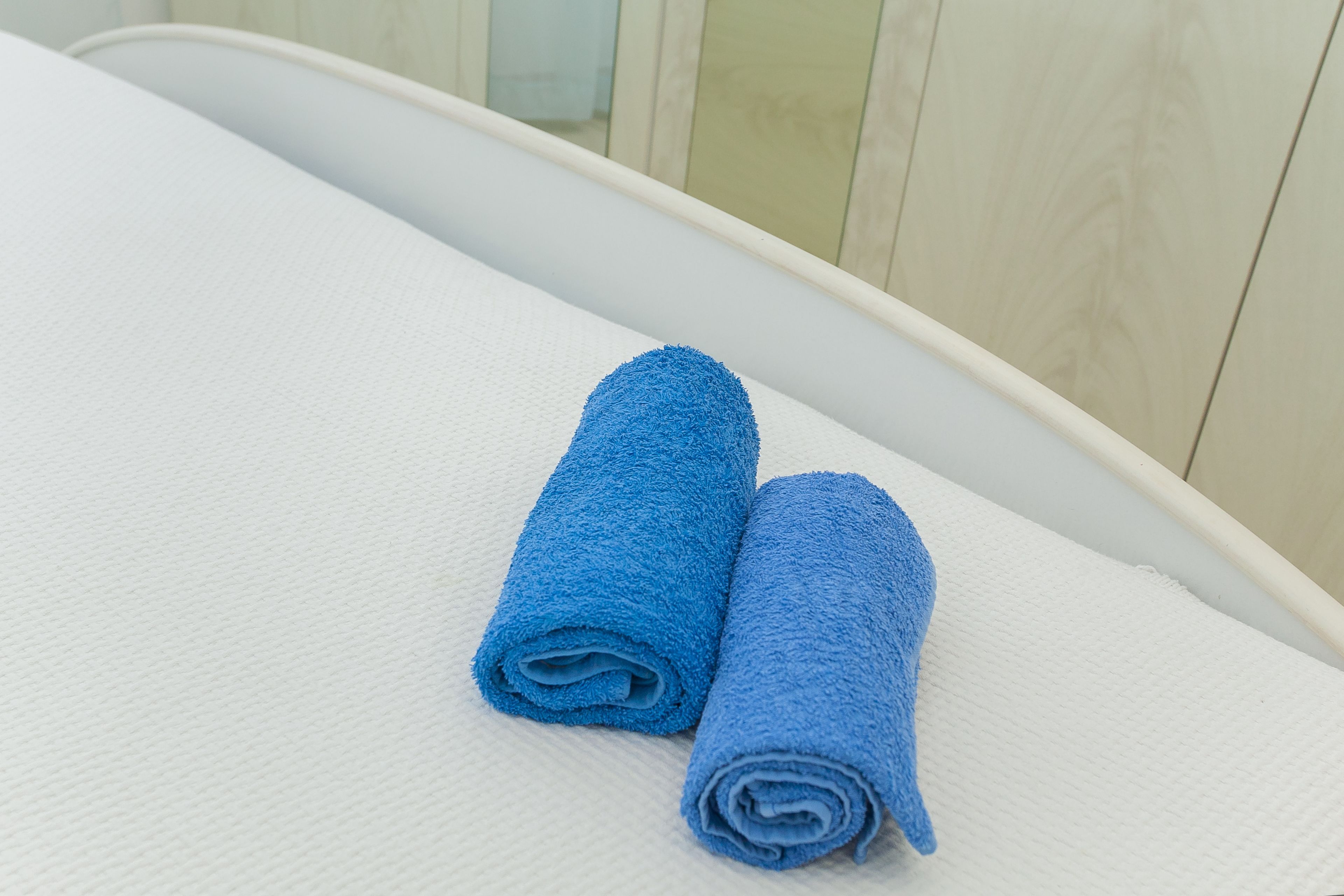Dos toallas azules enrolladas sobre una superficie blanca con textura, posiblemente una cama o sofá, en una habitación con paneles de madera clara.