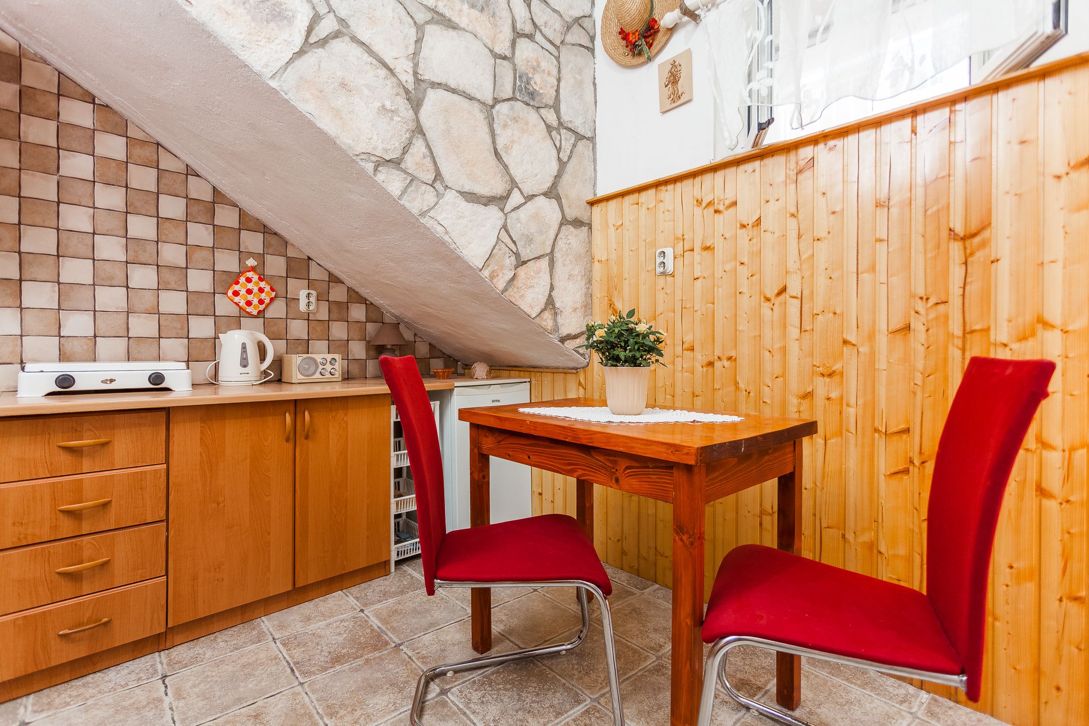 Une cuisine avec un mur en pierre, des armoires en bois, un pare-feu en carrelage, une table en bois avec deux chaises rouges et une petite plante en pot.