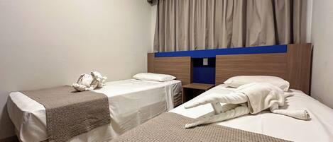 A hotel room with two single beds, a wooden headboard, a window with curtains, and a small table between the beds.