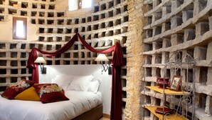 A bedroom with a stone wall, a bed with white bedding, red and yellow pillows, and a metal shelf with decorative items.